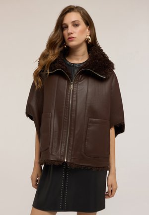 Mujer vestida con una chaqueta de cuero de piel de oveja de color marrón oscuro y gran collar sobre un vestido negro con tachuelas, contra un fondo liso.