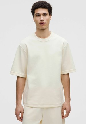 Giovane uomo con capelli corti e ricci che indossa una felpa ampia a maniche corte color crema e pantaloni abbinati, su uno sfondo semplice.