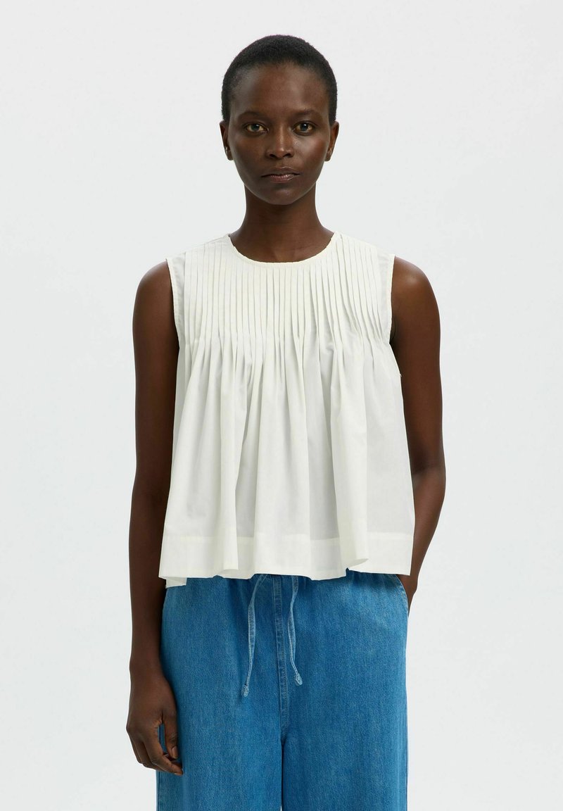 Femme aux cheveux courts portant un chemisier blanc plissé sans manches et un pantalon bleu ample taille haute, debout devant un fond uni.