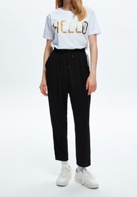 Vit T-shirt med guldtexten "HELLO", kombinerad med svarta, löstsittande byxor och vita sneakers. Enkel design, avslappnad passform och casual stil.