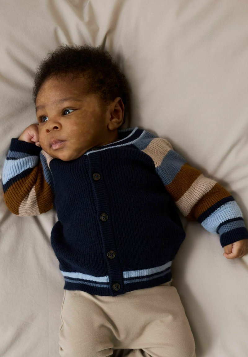 Blue and navy cardigan with striped long sleeves, featuring buttons and ribbed texture. Light tan bottom garment on a neutral background.