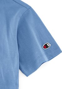 Camiseta de algodón azul de manga corta con cuello redondo y un logo bordado en la manga izquierda; textura suave y diseño casual.