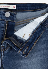 Supersmala jeans i mörkblå denim med dragkedja, kopparstickningar och en remsa av ljusblått tyg synlig inuti midjebandet.