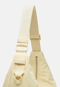 Helle beigefarbene Tasche mit strukturiertem Tragegurt, verstellbarem Schnallenverschluss und einem Ring in Goldton. An der Basis befindet sich ein Reißverschlussfach.