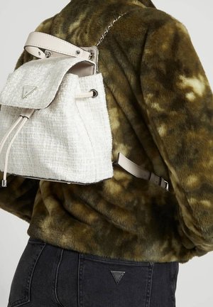 Witte tweed rugzak met trekkoorden, beige lederen accenten en zilverkleurige metalen onderdelen, gedragen over een groen-bruin fleecejack met patroon.