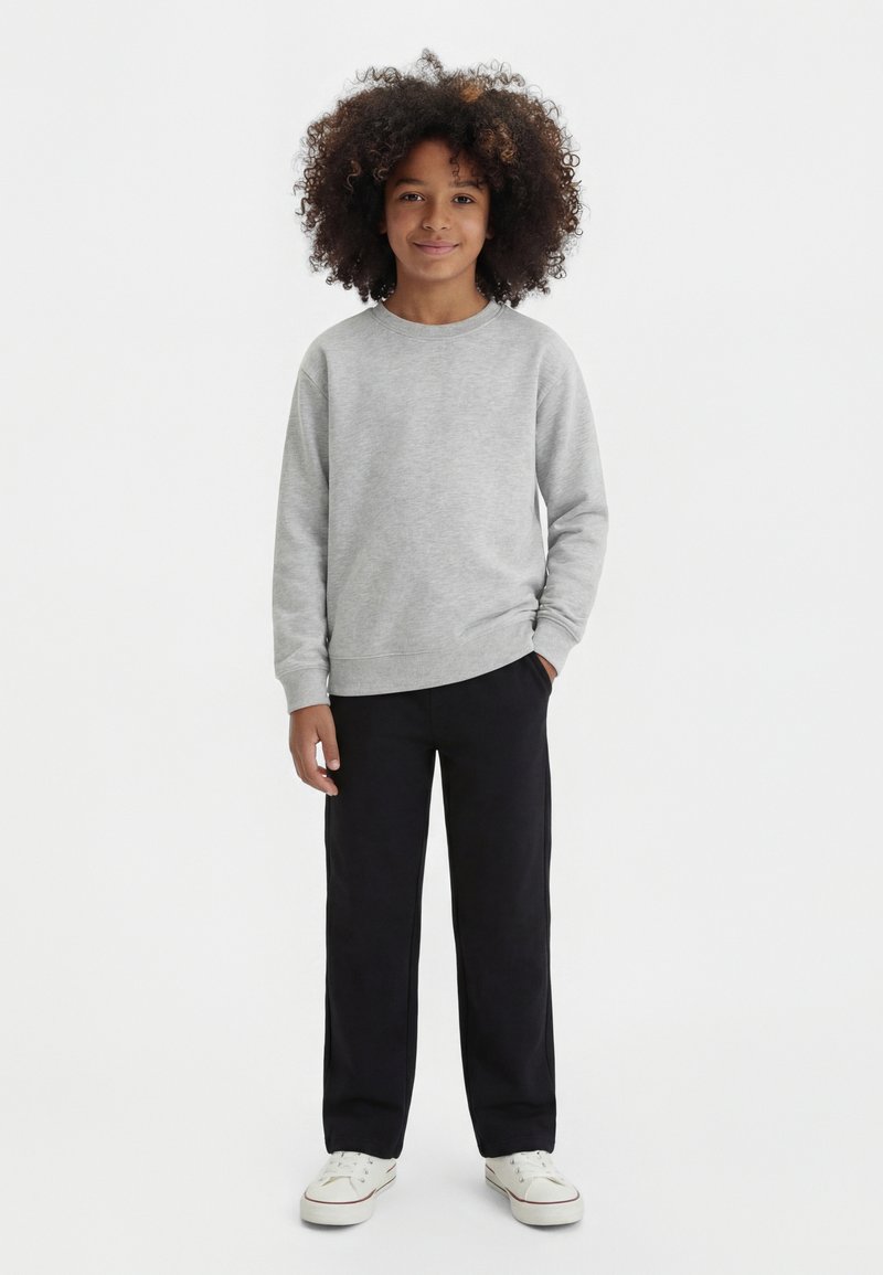 Dijete kovrčave kose u sivom džemperu, crnim hlačama i bijelim tenisicama, stoji s jednom rukom u džepu na bijeloj pozadini.