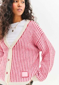 Červený a bílý proužkovaný svetr na knoflíky s výstřihem do V a texturovaným pletením. Má jednobílý lem a malou logovou nášivku.