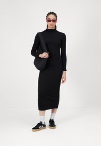 Μαύρο ριμπ πλεκτό φόρεμα με ψηλό λαιμό, μακριά μανίκια; συνδυασμένο με μια μαύρη καπιτονέ τσάντα ώμου και μαύρες αθλητικές μπότες με λευκές λεπτομέρειες.