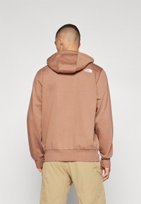 Brauner Kapuzenpullover mit Zugband und gerippten Bündchen. Hat ein kleines weißes Logo auf dem Rücken. Sweatshirtmaterial, lockere Passform.