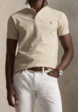 Mand iført beige poloshirt med lille logo, hvide bukser, brun bælte og brun læderarmbåndsur med mønstret urskive.