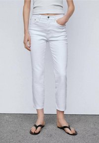 Jeans de mezclilla blanca con un diseño ajustado, estilo de cinco bolsillos y costuras mínimas. Combinados con chanclas negras, de pie sobre una superficie gris.