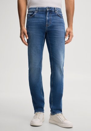 Mann trägt mittelblaue Slim-Fit-Jeans und weiße Sneaker, steht vor einem schlichten hellen Hintergrund.