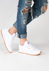 Weiße Sportsneaker aus glattem Leder mit orangefarbener Gummisohle, runder Zehenpartie und Logo auf der Zunge. Getragen mit ausgebeulten Jeans.