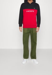 Röd och svart huvtröja med "LACOSTE"-logotyp, kombinerad med olivgröna byxor och vita sneakers, med en avslappnad passform och fickor.