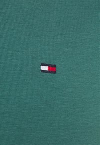 T-shirt in cotone verde con un piccolo logo ricamato in rosso, bianco e blu, posizionato sul lato sinistro vicino all'orlo.