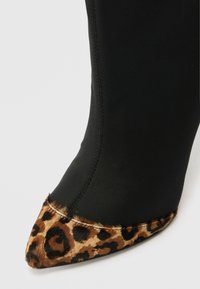 Botte en tissu noir avec un bout en fourrure léopard pointu. La botte présente un design élégant et des coutures visibles le long des seams.