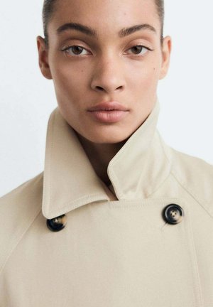Manteau beige avec un col haut, design à double boutonnage et boutons noirs. Texture lisse, style minimaliste.