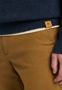 Maglione a coste navy sovrapposto a una maglietta crema; abbinato a pantaloni di tessuto marrone. Presenta un piccolo etichetta in pelle Timberland all'orlo del maglione.