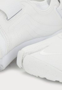 White athletic shoes with textured mesh uppers, Velcro strap, and solid rubber soles featuring subtle speckles and logo detail.