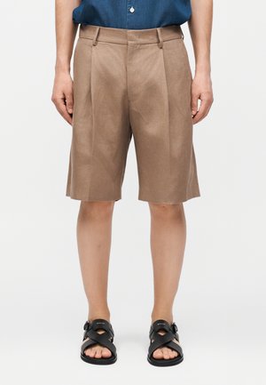 Beżowe plisowane krótkie spodenki do kolan noszone z czarnymi sandałami na klamrę i niebieską koszulą wpuszczoną do środka, widok z przodu na prostym tle.