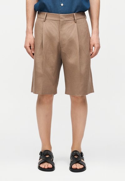 Short beige plissé mi-genou porté avec des sandales noires à boucle et une chemise bleue rentrée, vue de face sur fond uni.