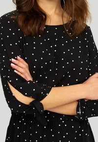 Robe noire à pois avec un col rond, des manches trois-quarts avec des accents noués, et un design à taille cintrée.