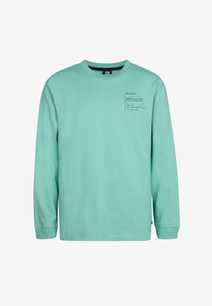 Langærmet mintgrøn t-shirt med et lille grafisk motiv på brystet, parret med grå jeans og hvide sneakers med sorte akcenter.