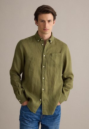 Joven con una camisa verde oliva abotonada con bolsillo en el pecho y vaqueros azules, de pie frente a un fondo beige liso.