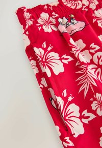 Tissu rouge avec grandes fleurs d'hibiscus blanches et feuilles vertes, avec une taille élastique et des détails sur les coutures latérales.