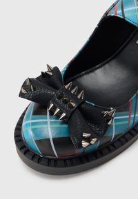 Krupni plan crne cipela s plavim kariranim uzorkom, sa crnim mašnom ukrašenom srebrnim metalnim bodljama na vrhu.