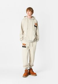 Conjunto de sudadera beige y pantalones de chándal con detalles de bolsillo bordados de oso. Textura suave, corte relajado, acompañado de botas de color tostado con cordones.