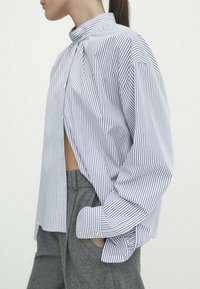 Chemise rayée à boutons bleue et blanche, avec un col haut, une coupe oversized et des manches bouffantes. Associée à un pantalon gris taille haute.