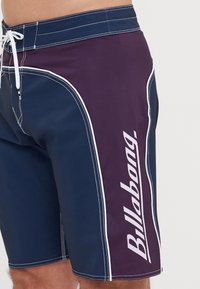 Marina blå och lila badshorts med vit sömnad. Har en sidologotyp och dragsko för stängning. Tillverkade av ett mjukt, snabbtorkande material.