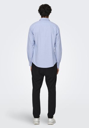 Camisa de botón azul claro con mangas largas, dobladillo curvado y botones blancos; combinada con pantalones negros y zapatillas blancas, vista desde atrás.