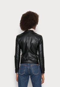 Svart läderjacka med en figursydd design, med en snedkrage och dragkedjedetaljer på ärmarna, bärs över en ljusfärgad tröja och blå jeans.