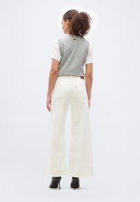 Γυναίκα με κατσαρά μαλλιά, που φορά γκρι αμάνικο πουλόβερ, λευκά παντελόνια με φαρδύ πόδι και μαύρες μπότες με ψηλό τακούνι, στέκεται με την πλάτη προς το θεατή σε λευκό φόντο.