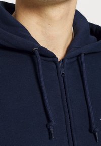 Námorně modrá zipová mikina vyrobená z měkkého materiálu, s předním zipem, kapucí na šňůrku a texturovanými švy.