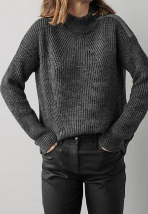 Pull en maille grise avec texture côtelée, col montant et épaules tombantes, associé à un pantalon noir ajusté.