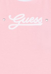 Gaiši rozā T-krekls ar baltu slīprakstā rakstītu "Guess" logo, kurā abās pusēs ir mazi sudraba sirds akcenti. Mīksta auduma tekstūra.