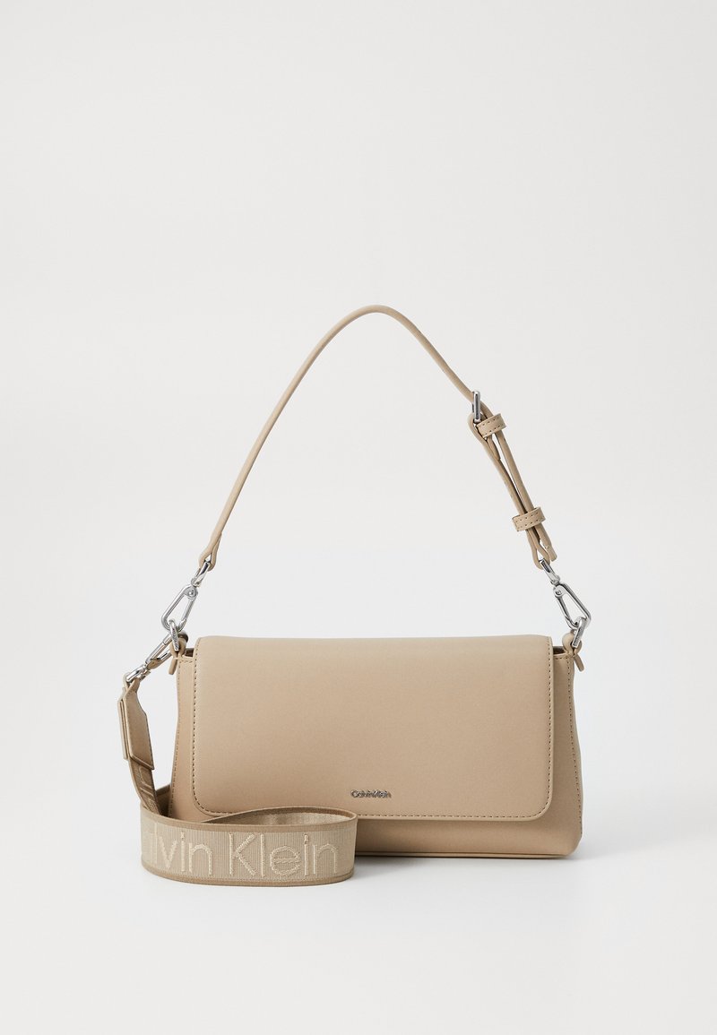 Beige synteettinen käsilaukku, jossa on sileä pinta, ylhäällä läppäkiinnitys ja irrotettava hihna, jossa on "Calvin Klein" vaaleammalla sävyllä.