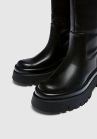 Botas de cuero negro con una suela de goma gruesa, punta redondeada y un acabado suave. Presentan costuras mínimas y sin herrajes visibles.