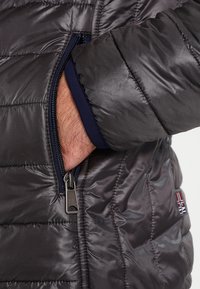 Graue isolierte Jacke mit einer Reißverschlusstasche. Sie hat eine glatte, glänzende Textur, ein gestepptes Muster und einen kontrastierenden marineblauen Innenfutter. Reißverschlussbeschläge sichtbar.