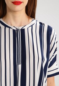 Blouse à rayures bleu marine et blanches en tissu léger. Elle présente un col rond avec une patte de boutonnage partielle et des manches courtes.