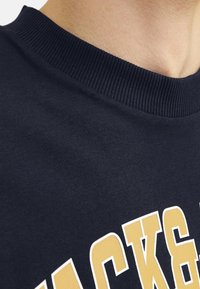Navy cotton sweatshirt features a ribbed crew neckline and bold yellow lettering. The fabric has a smooth texture with minimal detailing.
