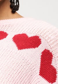 Κοντινή λήψη ροζ πλεκτού πουλόβερ με τρία μεγάλα κόκκινα μοτίβα καρδιάς στον ώμο και το πάνω μέρος της πλάτης.