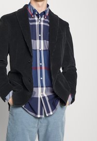 Zwarte corduroy blazer met twee knopen, gedragen over een blauw-wit geruit overhemd. Blauwe gestreepte broek maakt de outfit compleet.