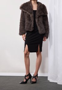 Veste courte en fausse fourrure marron avec un large col, portée sur une robe noire ajustée avec des fentes latérales. Chaussures à talons hauts noires avec des brides cloutées.