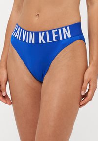 Blauwe bikinionderkant met een brede witte Calvin Klein logo band. De stof lijkt glad en rekbaar, met een lage taille.