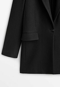 Manteau en laine noire à manches longues, avec une fermeture à un bouton noir et un rabat de poche sur le côté droit.