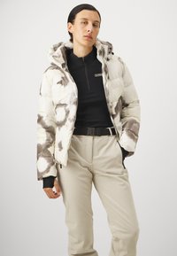 Gepolsterte Jacke mit Kapuze, die ein schwarz-weißes abstraktes Muster aufweist. Getragen über einem schwarzen Reißverschlussoberteil und beigen gerippten Hosen.